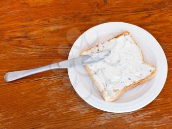 top view of sandwich from toast and cottagecheese with herbs on white plate, table knife on wooden table