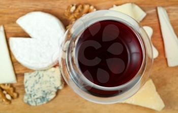 top view of glass of red wine and various cheeses on wooden plate close up