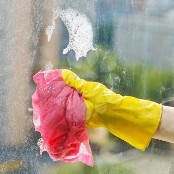 hand in yellow rubber glove cleaning window glass by soap suds water