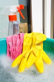 home window washer set - yellow rubber glove, wet rag, spray glass cleaner bottle, foamy water in green basin on windowsill