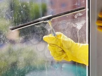 hand in yellow glove cleans home window glass by squeegee in spring day
