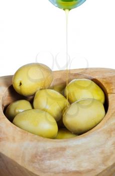 olive oil in thin stream flows from bottle on green olives in wooden bowl close up isolated on white background