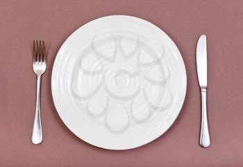 top view of empty white plate with fork and knife set on brown background