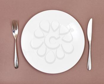 top view of empty white porcelain plate with fork and knife set on brown background