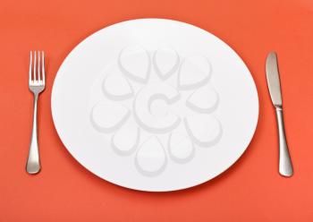 above view of empty white porcelain plate with fork and knife set on red background