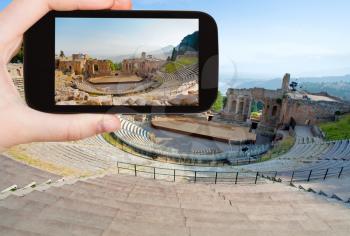 travel concept - tourist taking photo of ancient amphitheater Teatro Greco, Taormina, Sicily on mobile gadget, Italy