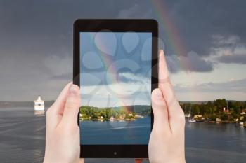travel concept - tourist takes picture of rainbow over small village on Baltic sea coast on smartphone, Sweden