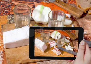 photographing food concept - tourist takes picture of russian appetizers two glass with vodka, salty lard and fresh onion on table in rural home on smartphone,