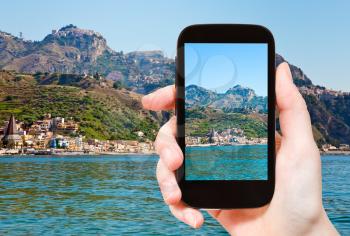 travel concept - tourist takes picture of Taormina - popular tourist resort on mountain and Gardini Naxos beach, Sicily on smartphone