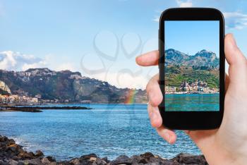 travel concept - tourist takes picture of town Taormina and Giardini Naxos beach, Sicily on smartphone