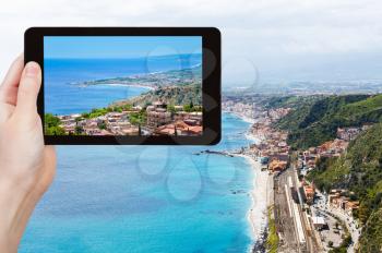 travel concept - tourist takes picture of Ionian Sea coastline and Giardini Naxos town from Taormina city, Sicily, Italy on tablet pc