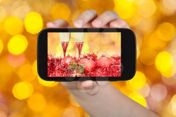 man takes photo of Christmas still life - two glasses of champagne at red Xmas decorations with yellow and brown blurred Christmas lights background