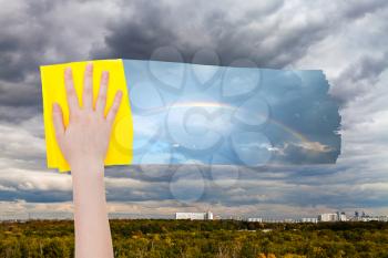weather concept - hand deletes gray rainy clouds over city by yellow cloth from image and rainbow is appearing