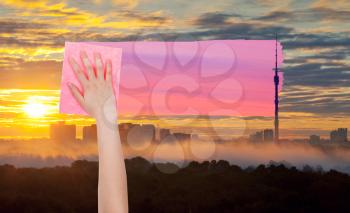 weather concept - hand deletes yellow sunrise sky over city by pink cloth from image and pink sunset sky is appearing