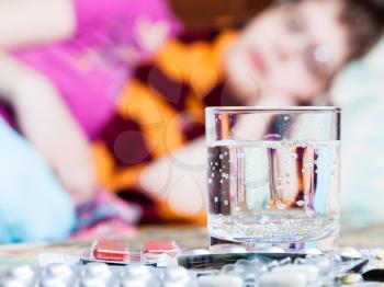 glass with water and pile of pills on table close up and sick woman with scarf around her neck on sofa in bed-sitting room on background