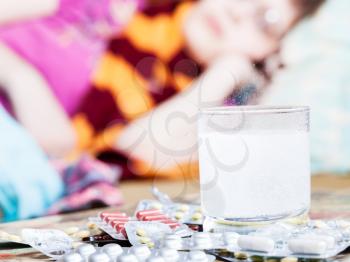 drug dissolves in water and pills on table close up and sick woman with scarf around her neck on sofa in living room on background