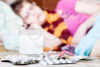 dissolving drug in glass with water and pile of pills on table close up and sick woman with scarf around her neck on sofa in bed-sitting room on background