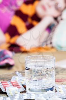 glass with medicament and pile of pills on table close up and sick woman with scarf around her neck on sofa in bed-sitting room on background