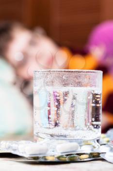 glass with dissolved medicament in and pills on table close up and sick woman with scarf around her neck on sofa in living room on background