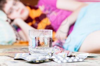 glass with water and pills close up and sick girl with scarf around her neck on sofa in living room on background