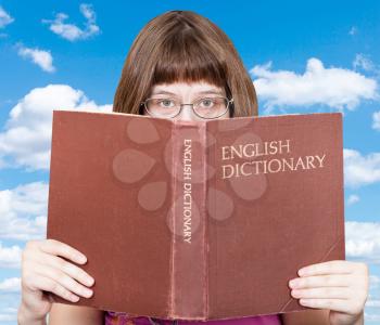 girl with spectacles looks over English Dictionary book with white clouds in blue sky on background