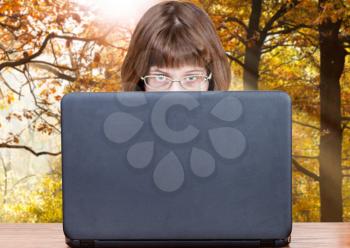 girl with spectacles looks over cover of open laptop with autumn scenery background