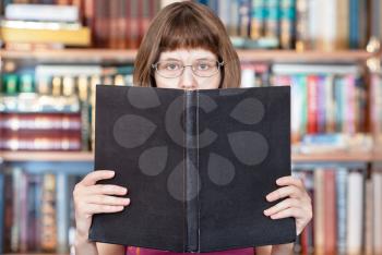 girl with glasses reads big book with blank cover and book shelves on background