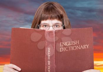 girl with spectacles looks over English Dictionary book and red yellow sunset sky on background