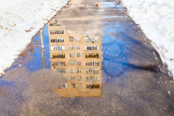reflection of apartment house in puddle of melting snow on urban footpath in sunny spring day