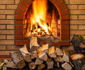 stack of firewood and fire in indoor brick fireplace in country cottage