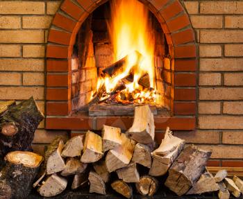 pile of firewood and fire in indoor brick fireplace in country cottage