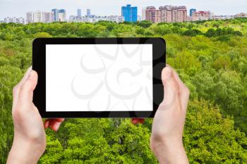 season concept - tourist photographs green trees in summer forest and urban houses on horizon on tablet pc with cut out screen with blank place for advertising