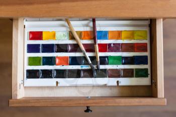 above view of watercolor paints set and painting brushes in open drawer of nightstand