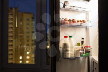 Open door of home fridge with dairy products and view of city through window in night