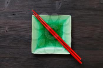 top view of one green square saucer with red chopsticks on dark brown table