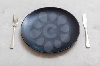 food concept - black plate with knife, spoon on gray concrete surface