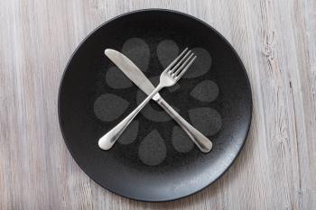 food concept - top view of black plate with crossing knife, spoon on gray brown table