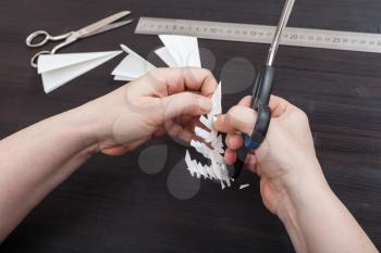 top view of hand with scissors cut a figure of snowflake from folded sheet of paper on dark brown wooden table