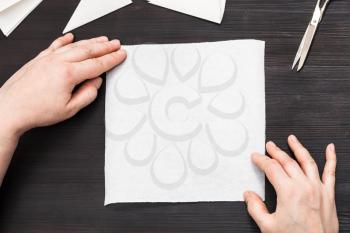 top view of hands hold blank piece of paper before folding to make figure on dark brown wooden table