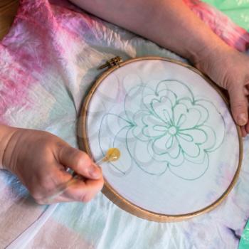 batik painting workshop - artist draws contour of flower on batik on white silk canvas clamped in the hoop