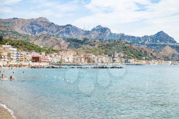 travel to Sicily, Italy - people in Ionian sea near waterfront of Giardini Naxos town and view of Taormina city on cape in summer