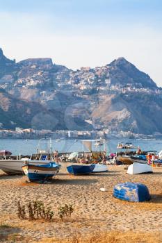 travel to Sicily, Italy - boats on beach in old port in Giardini Naxos town and view of Taormina city on cape in summer