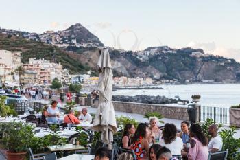 GIARDINI NAXOS, ITALY - JULY 2, 2017: people in sidewalk restaurant on waterfront in Giardini-Naxos town summer evening. Giardini Naxos is seaside resort on Ionian Sea coast since the 19