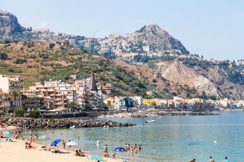 GIARDINI NAXOS, ITALY - JUNE 27, 2017: people on urban beach in Giardini-Naxos town and view of Taormina city on cape. Giardini Naxos is seaside resort on Ionian Sea coast since the 1970s