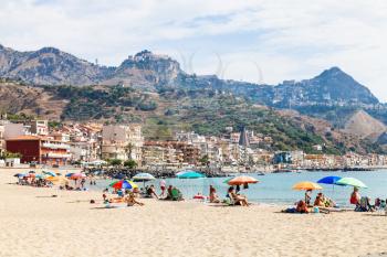 GIARDINI NAXOS, ITALY - JUNE 28, 2017: people on sand beach in Giardini-Naxos town and view of Taormina city on cape. Giardini Naxos is seaside resort on Ionian Sea coast since the 1970s