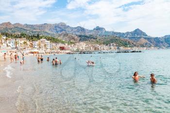 GIARDINI NAXOS, ITALY - JUNE 28, 2017: people in sea near waterfront of Giardini-Naxos town and view of Taormina city on cape. Giardini Naxos is seaside resort on Ionian Sea coast since the 1970s