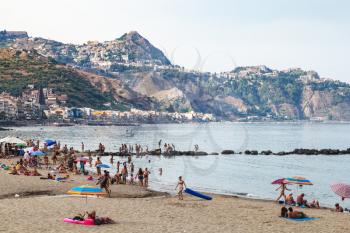 GIARDINI NAXOS, ITALY - JUNE 28, 2017: people on urban beach in Giardini-Naxos town in evening and view of Taormina city on cape. Giardini Naxos is seaside resort on Ionian Sea coast since the 1970s