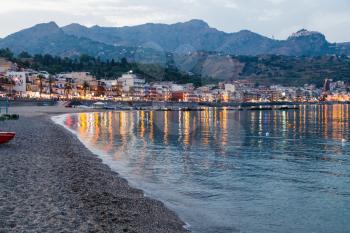 GIARDINI NAXOS, ITALY - JUNE 28, 2017: people on beach in Giardini-Naxos town view of Taormina city on cape in summer evening. Giardini Naxos is seaside resort on Ionian Sea coast since the 1970s