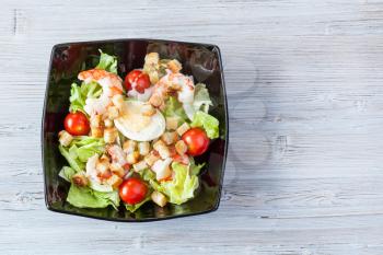 top view of Caesar salad with prawns in black bowl on gray wooden table with copyspace