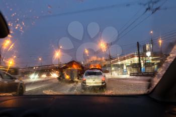 view of car traffic and street sweeping machines through wet windscreen while driving car on road in Moscow city in winter evening in snow